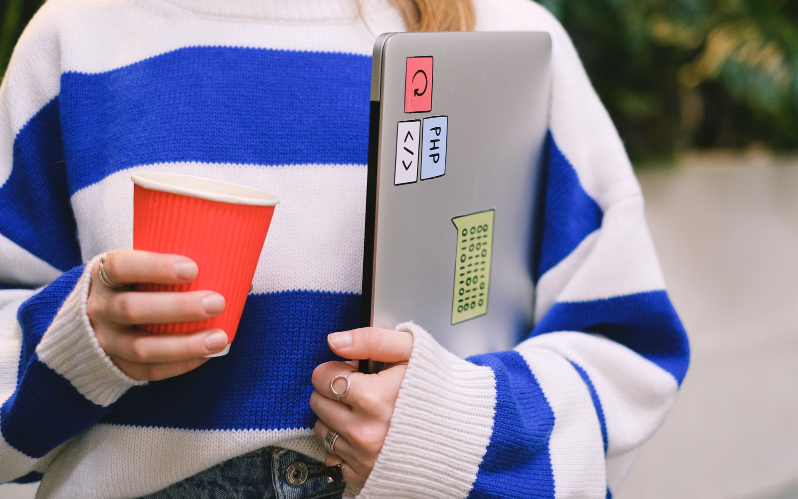 Person wearing striped sweater holding laptop with stickers and a red coffee cup indoors.