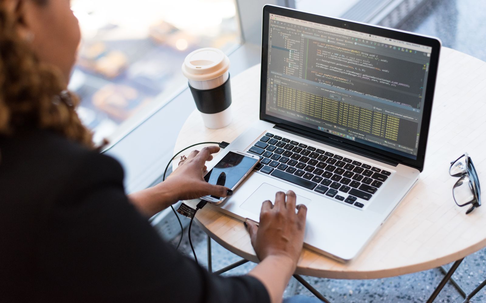 Black woman programming on a laptop with coffee, smartphone, and glasses on a desk in an office.