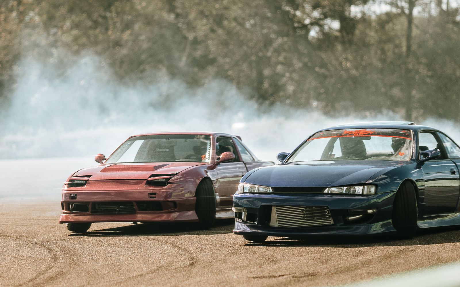 Two sports cars drift around a track emitting smoke, showcasing speed and precision.
