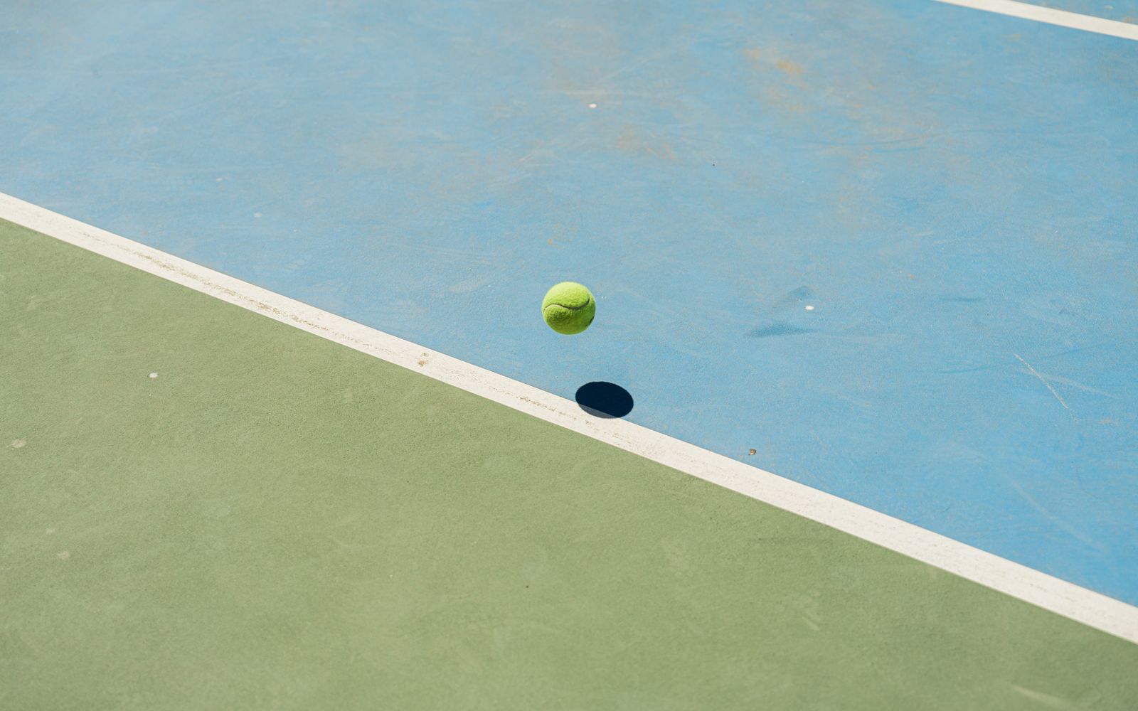 A tennis ball bounces midair creating a shadow on a blue and green tennis court.