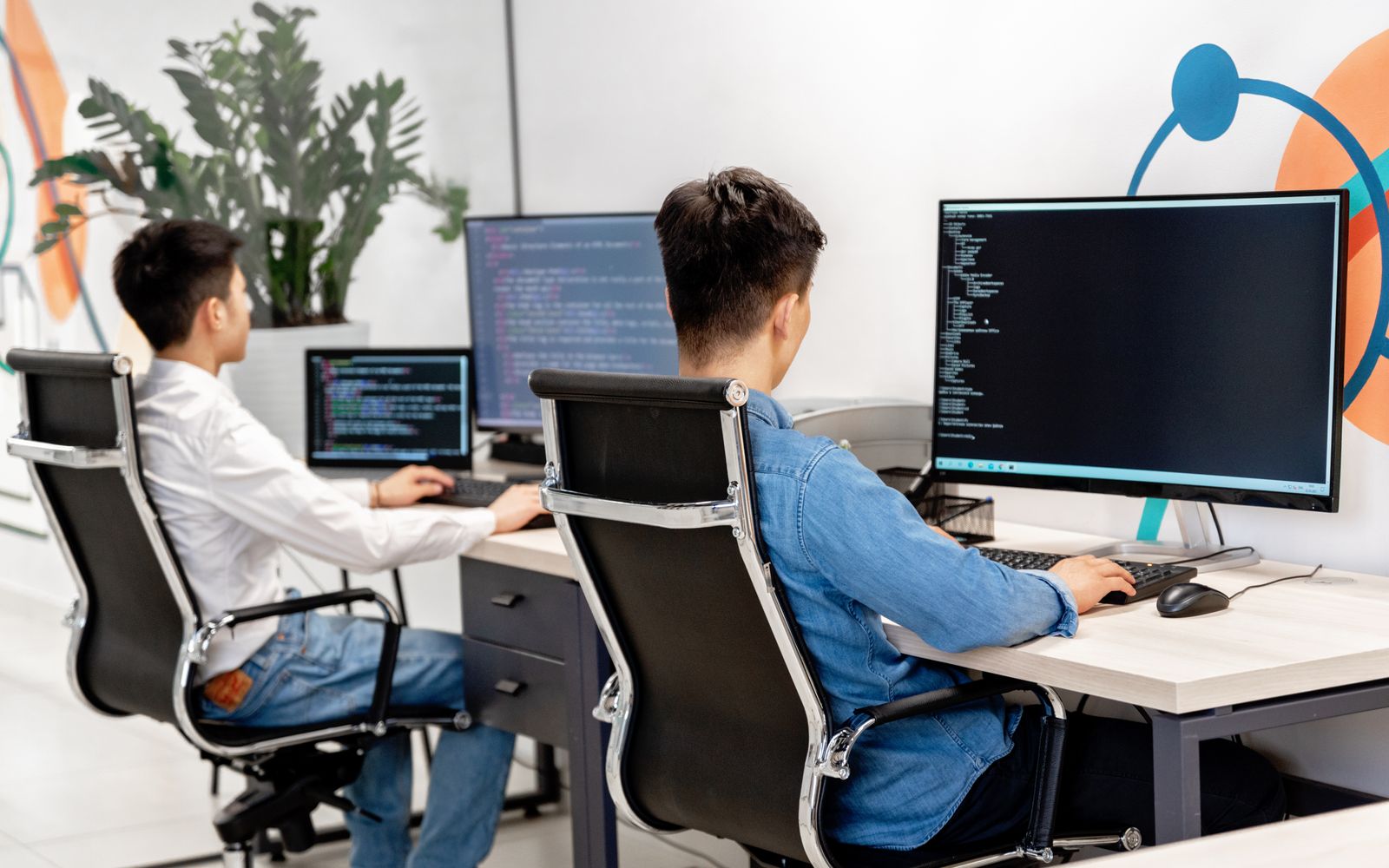 Two male developers at desks programming in a modern office workspace with large monitors.