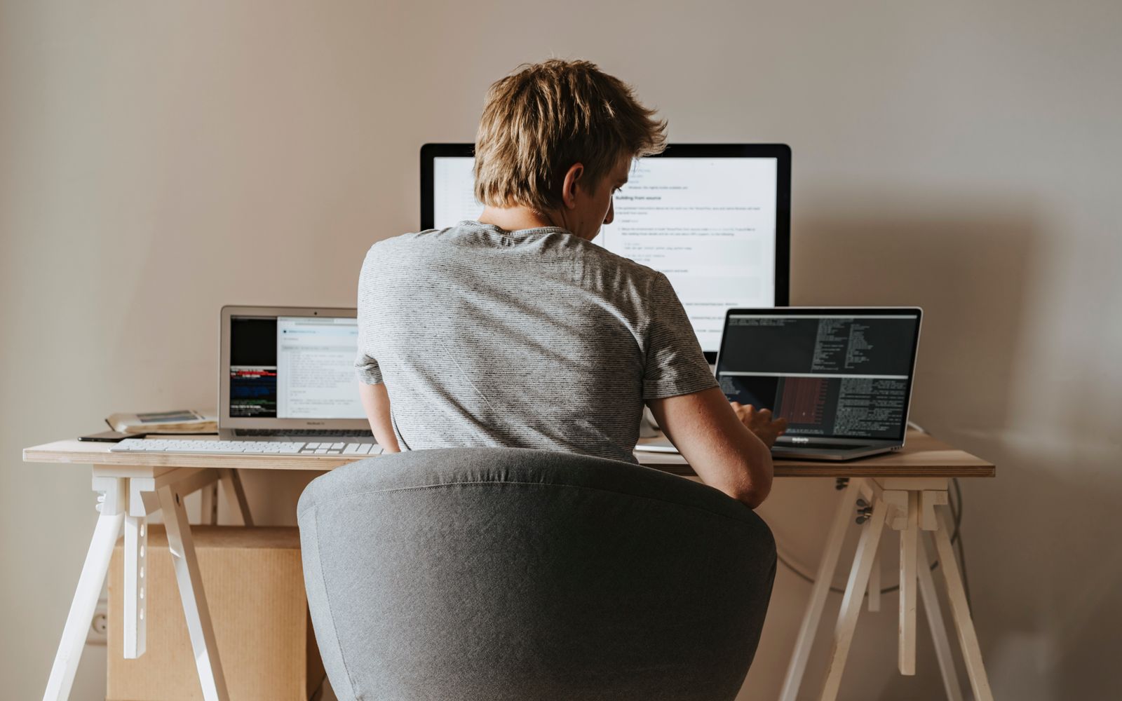 Young man coding at home office with multiple laptops, representing remote work and technology.