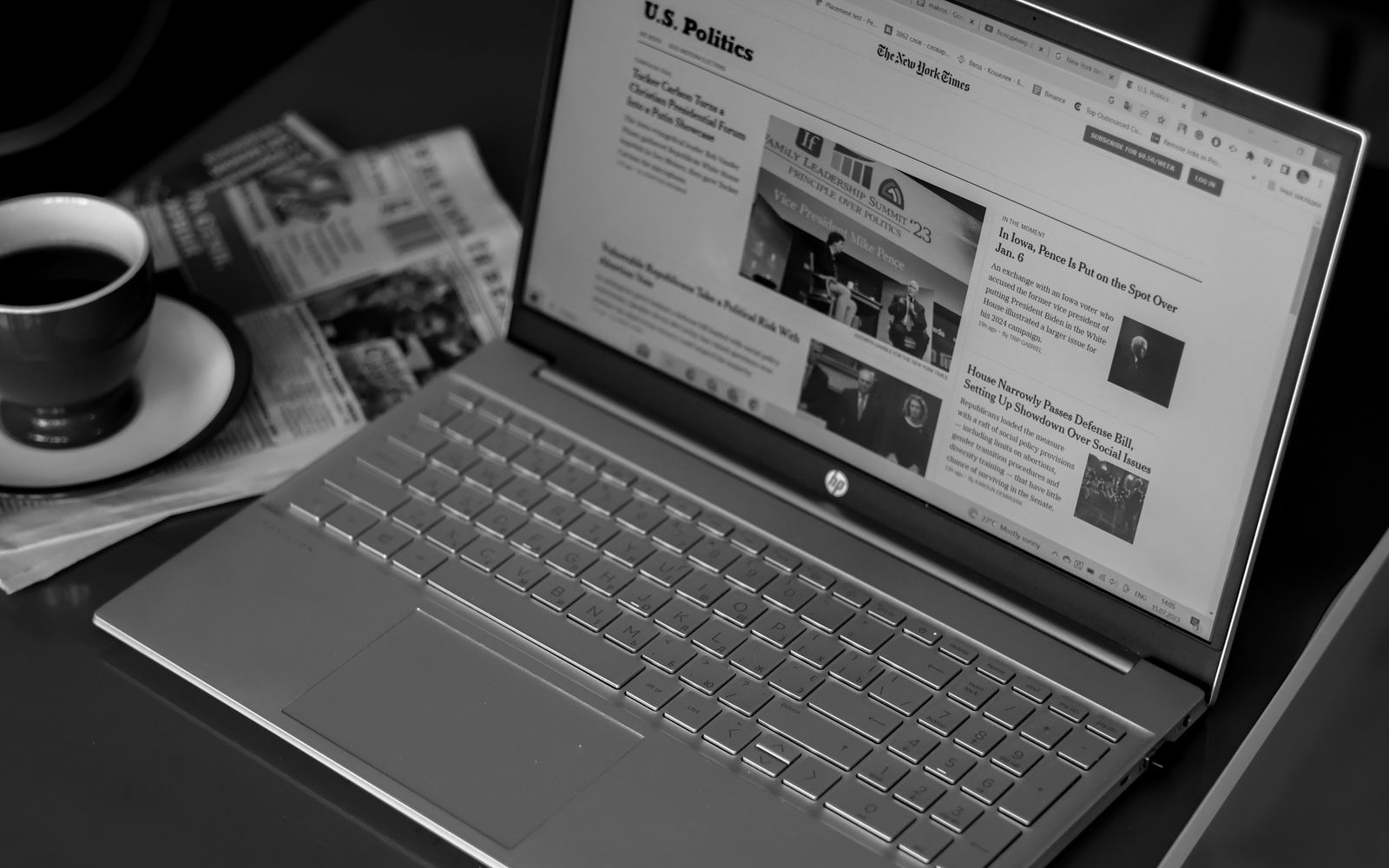 Black and white image of a laptop displaying news articles, accompanied by a cup of coffee and newspapers.