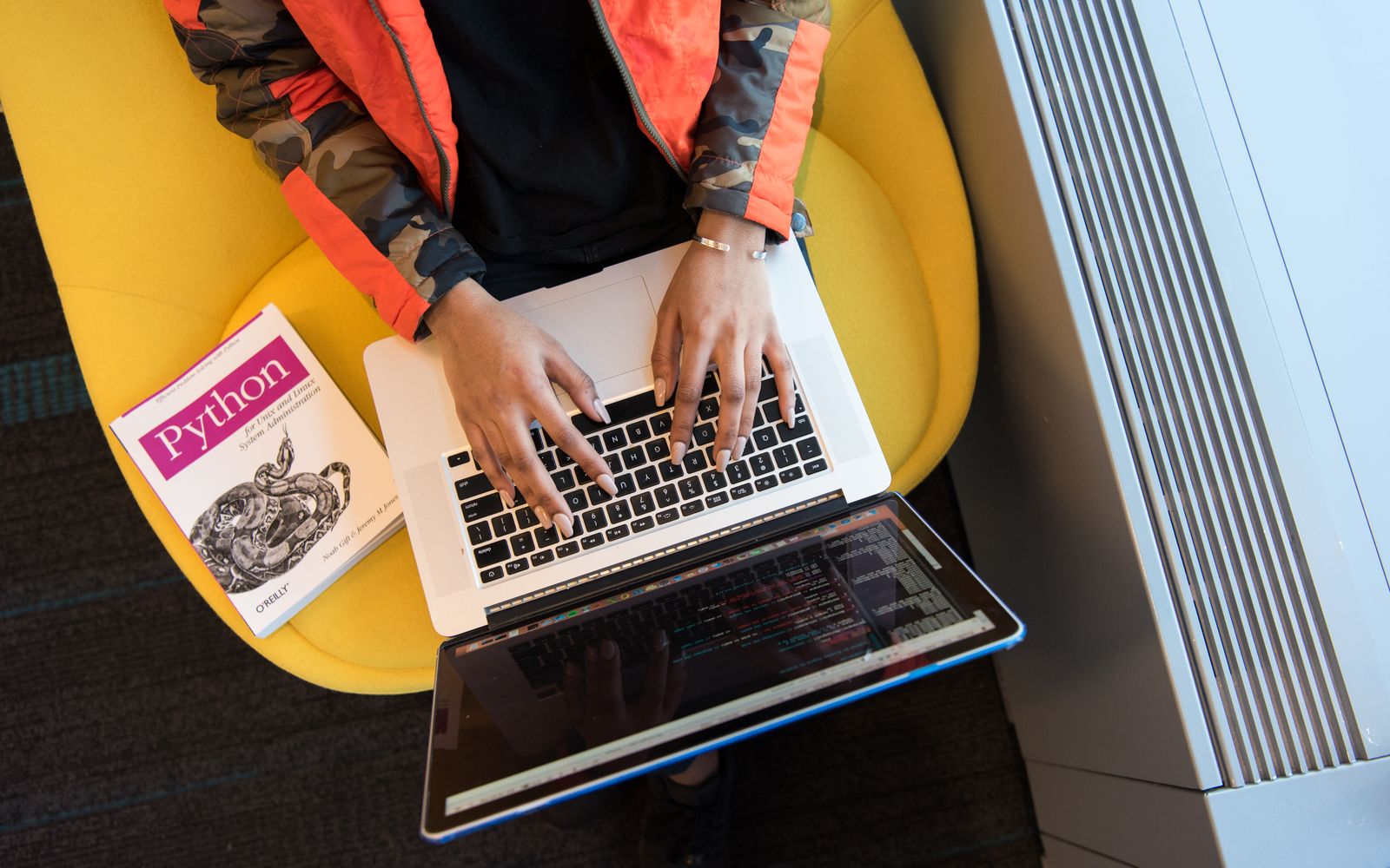 High-angle view of woman coding on a laptop, with a Python book nearby. Ideal for programming and tech content.