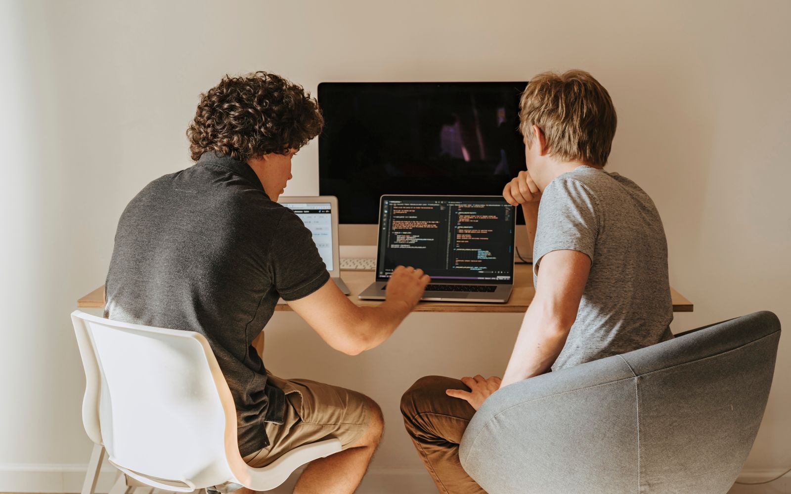 Two male developers working on laptops indoors, discussing code.