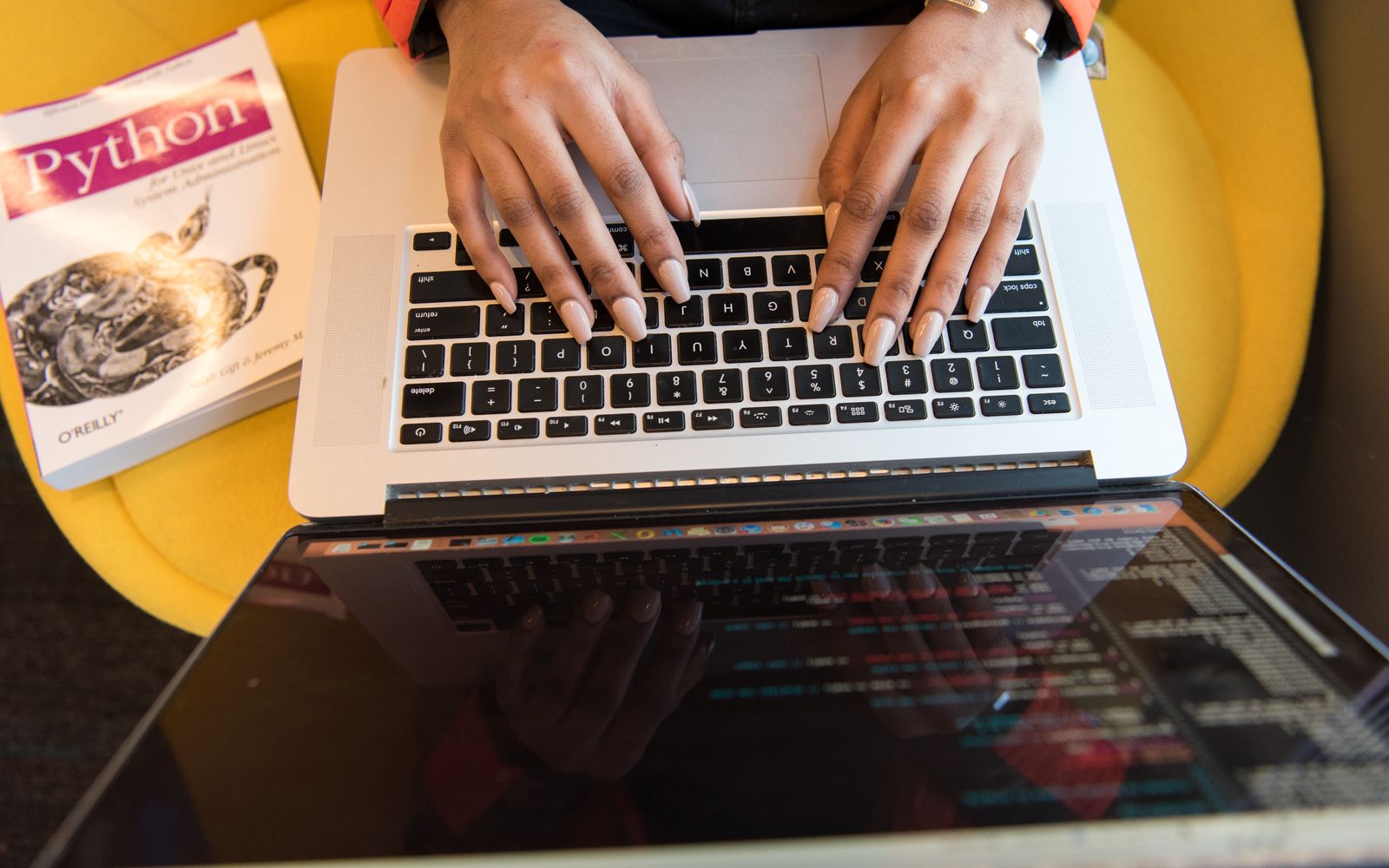 Close-up of hands typing on a laptop keyboard, Python book in sight, coding in progress.