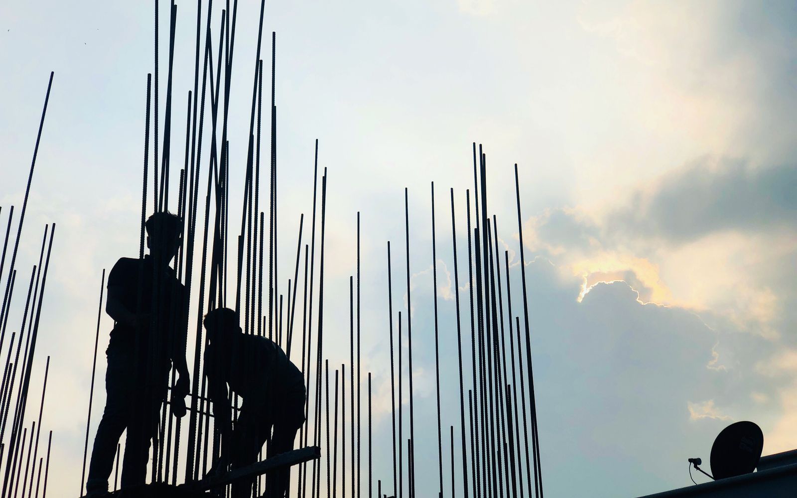 Silhouetted construction workers on site at dawn in Dhaka, Bangladesh, against a cloudy sky.