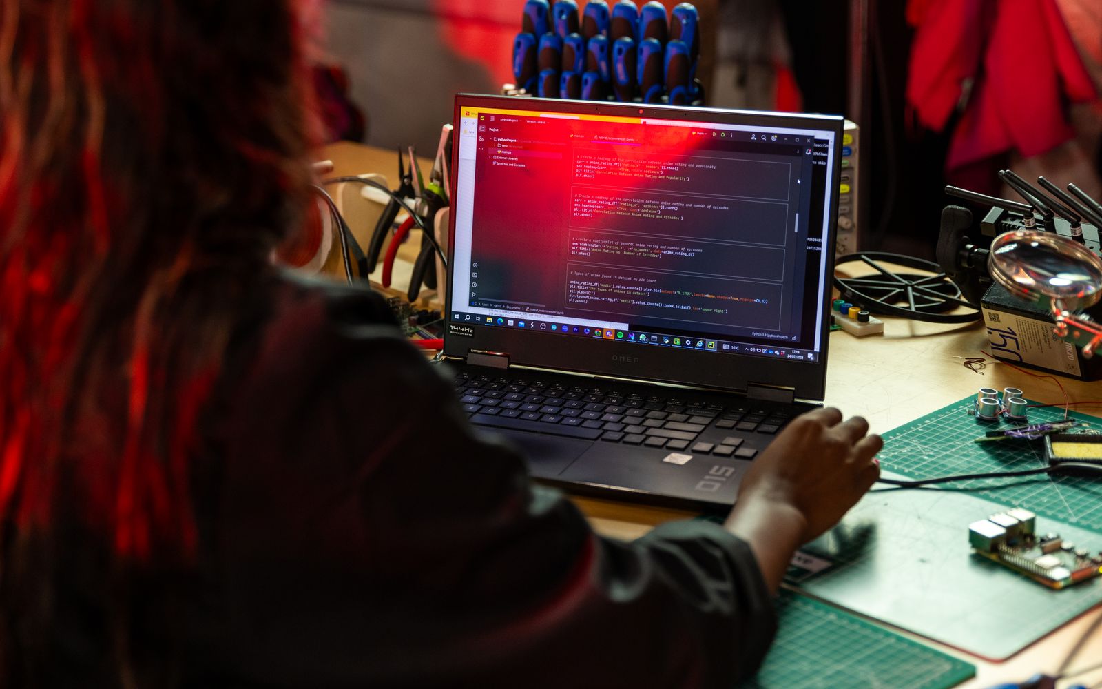 A software developer writing code on a laptop at a cluttered workbench with electronic tools.